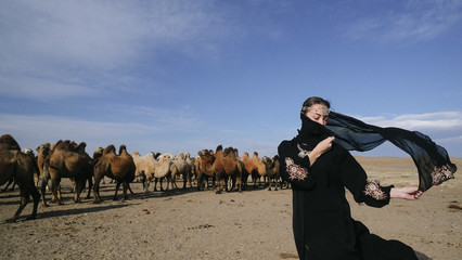 beautiful woman national eastern clothes black color,veil,hijab,abaya ornament her head,portrait steppes camels,strong wind,desert,wildlife,well,decoration,caucasian,slow ,sun