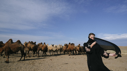 beautiful woman national eastern clothes black color,veil,hijab,abaya ornament her head,portrait steppes camels,strong wind,desert,wildlife,well,decoration,caucasian,slow ,sun