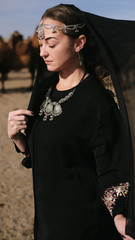 beautiful woman national eastern clothes black color,veil,hijab,abaya ornament her head,portrait steppes camels,strong wind,desert,wildlife,well,decoration,caucasian,slow ,sun