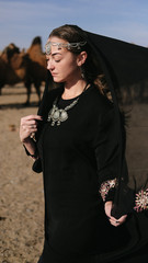 beautiful woman national eastern clothes black color,veil,hijab,abaya ornament her head,portrait steppes camels,strong wind,desert,wildlife,well,decoration,caucasian,slow ,sun