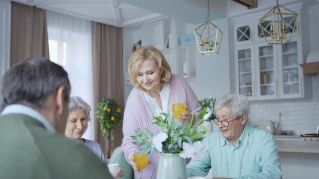 Tracking shot of caring senior woman bringing glasses of orange juice for her elderly friends sitting at table and having desserts at home