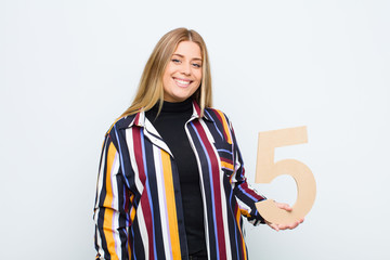 young pretty blonde woman excited, happy, joyful, holding a number 5.