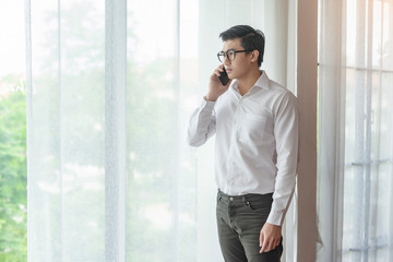 Asian male architecture holding a smartphone to his ears and calling or answering, standing straight looking outside of the window with a serious face, wearing a white long sleeve shirt with jeans...