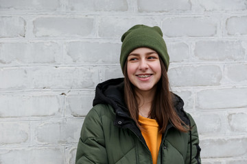 Portrait of a beautiful smiling girl with braces in a yellow sweater and khaki hat, which stands near a white brick wall. The concept of emotions and copy space