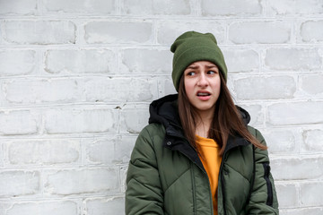 Portrait of a beautiful incredulous girl with braces in a yellow sweater and khaki hat that stands near a white brick wall. The concept of emotions.