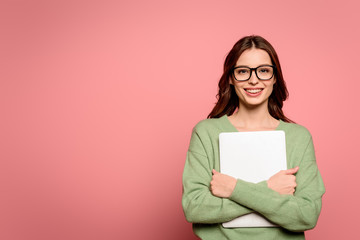 happy businesswoman in glasses holding closed laptop while smiling at camera on pink background