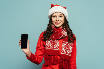 smiling girl in santa hat and red sweater showing smartphone with blank screen on blue background