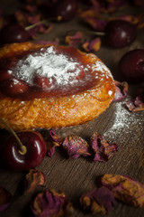 Cherry jam tart with raw cherries laid around and dried roses on wooden boards on black background.