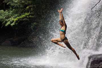 Young athletic woman jumping into water with raised hands. Background of many drops of waterfall and tropical forest