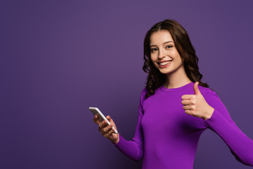 Naklejka premium cheerful girl holding smartphone and showing thumb up on purple background