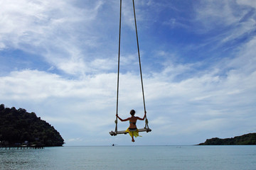 Young woman in bikini swinging on rope swing over ocean water on tropical beach. Blue sky, horizon, sea. View from back