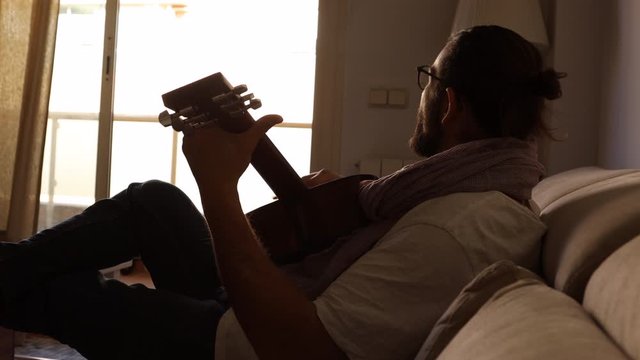 Young man musician with beard and long hair writing songs lying on the sofa of his apartment in his notebook