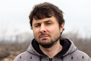 A handsome bearded man with disheveled hair squinted his eye in displeasure against a blurred background. Man in a jacket with a hood