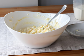 Mixing cottage cheese dough in a white bowl with a fork on a linen tablecloth on a wooden table. Ingredients - cottage cheese, butter, egg and sugar. The process of making curd bagels or croissants