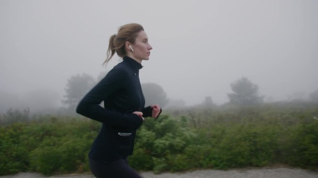 Lateral Tracking Cinematic Shot of Young Woman Wearing Headphones Running in Foggy Park. Female Runner Exercising Outdoors as Part of Her Morning Jogging Routine. Healthy Lifestyle and Fitness. Slow M
