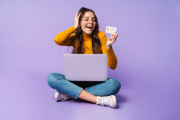 Image of young woman holding credit card and using laptop while sitting