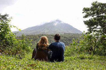 back of Young couple looking at Arenal volcano, sitting on green grass and enyojing nature, Costa Rica, Central America, la fortuna - obrazy, fototapety, plakaty