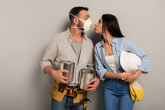 Couple Of Happy Manual Workers In Safety Mask Holding Paint Cans And Helmet And Kissing On Grey