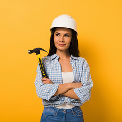 Cheerful workwoman in helmet holding hammer with crossed arms on yellow