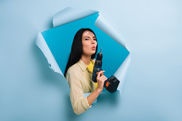 confident handywoman with dirty face in paint holding electric drill in torn paper, isolated on blue