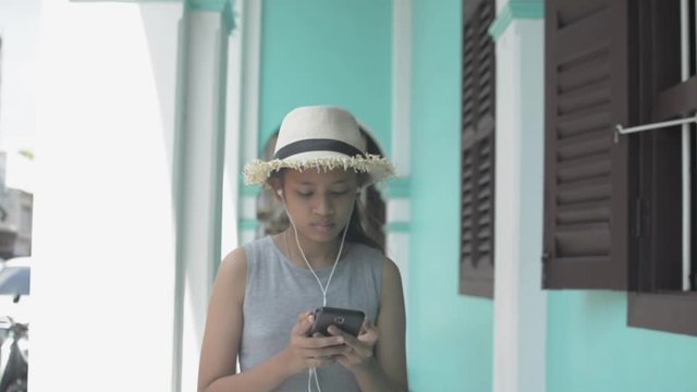 Asian cute girl in casual dress wearing straw hat using mobile phone and earphones walking leisurely in side arcade in chino portuguese style building, Phuket, Thailand. Slow motion.