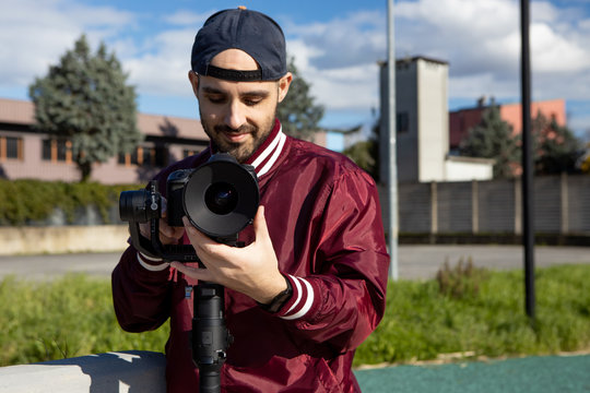 Caucasian young man with reverse cap and red jacket works outdoor with his camera on a gimbal stabiliser. Video operator focusing with his video camera. Cinema lens on DLSR camera. Medium angle shot