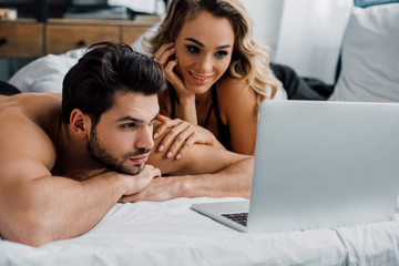 Handsome man looking at laptop near smiling woman on bed