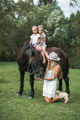 Happy family, mother with two children cute daughters riding a horse. Mother in white dress, hat and cowboy boots standing on the knees and feeding the horse. Family time on nature, horse riding