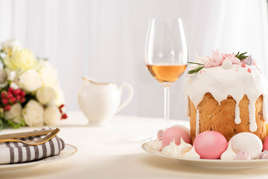 selective focus of delicious Easter cake decorated with meringue with pink and white eggs on plate near wine glass and flowers