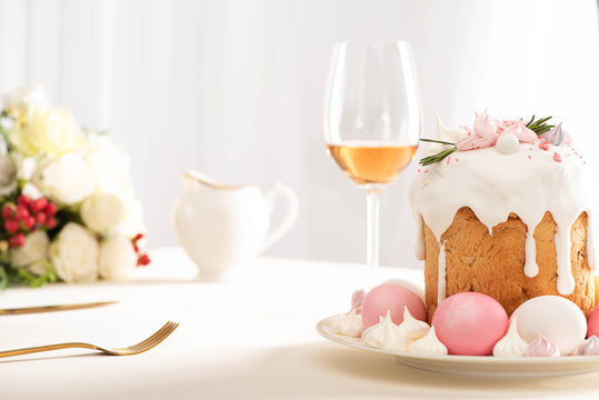 selective focus of delicious Easter cake decorated with meringue with pink and white eggs on plate near wine glass and flowers