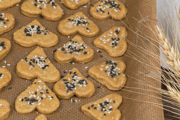 Gingerbread cookie hearts on baking paper on a baking sheet on a wooden table. The scene of making sweet homemade pastries with black and white sesame, sugar. Rustic style recipe