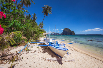 Fototapeta na wymiar Colored banca boat and vibrant flowers at Las cabanas beach. Surreal landscape in background. Exotic nature scenery in El Nido, Palawan, Philippines