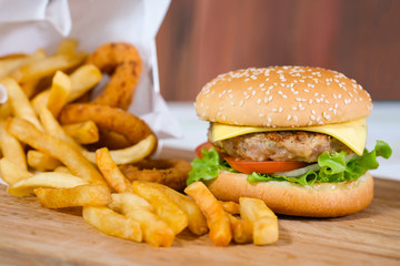 Close-up macro of delicious fresh homemade hamburger with cheese, pork, tomato, onion and vegetable on a wooden plate with french fries basket and onion ring.