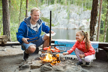 Man and his little daughter having barbecue in forest on rocky shore of lake, making a fire, grilling bread, vegetables and marshmallow. Family exploring Finland. Scandinavian summer landscape. 