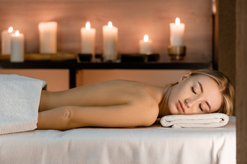 Obraz na płótnie Canvas Spa concept. Young beautiful slim girl lies on massage table in anticipation of massage on a background of blurry burning candles.Front view, copy space
