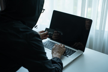 Rearside of man dressing black hood jacket holding credit card and checking password on labtop computer. hacker scammer concept - obrazy, fototapety, plakaty
