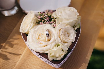 White roses in a paper box made in form of the heart with wedding rings, on a wooden table