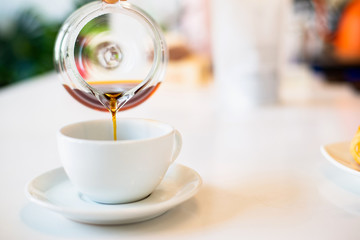 Barista is pouring hot coffee from the coffee pot into the white coffee on the wooden table in the afternoon.