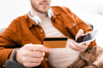Selective focus of 3d artist in headphones holding smartphone and credit card on couch on white background