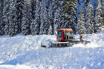 A snow removal and ski tracks making machine removes snow in a ski resort in winter. Snow-covered Forest on the hill is in the background.