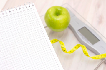 green apple, a measuring tape, scale and a notebook for writing on a light wooden background. preparation for the summer season and the beach, weight loss and sports concept