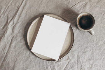 Cup of coffee and blank greeting card, invitation on ceramic plate. Moody breakfast table mockup...
