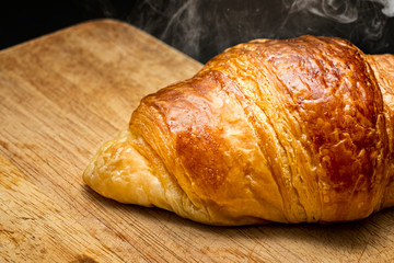 Close-up fresh new baking golden croissant  fragrant in warm light with thin smoke, on cutting board. Wooden texture.Concept of fresh new breakfast on black background.with clipping path.