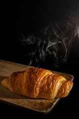 Fresh new baking croissant  fragrant in warm light with thin smoke, on grunge cutting board. Wooden texture.Concept of fresh new breakfast on black background.