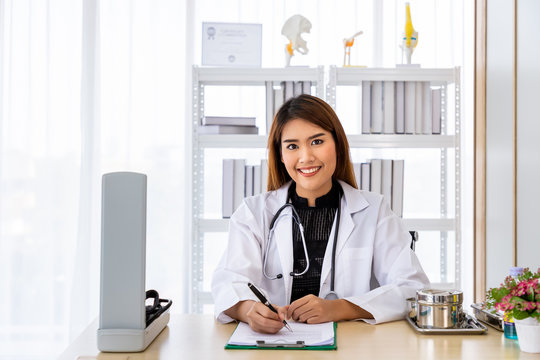 Portrait Of A Smiling Asian Medical Doctor Sitting At Desk In Office Clinic Hospital Wearing White Coat With Stethoscope, Looking At Camera