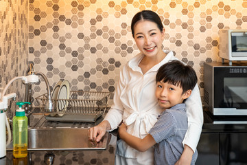 Beautiful young Asian boy son hugging woman mother in modern kitchen room, looking at camera