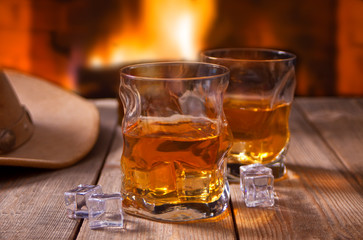 whiskey with ice on a wooden table with fireplace and cowboy hats on the background.