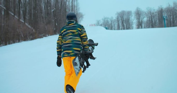 Snowboarder climbs the mountain in a ski suit and with a snowboard. Professional snowboarder is walking along the track, holding a snowboard in his hands. Beautiful winter landscape.