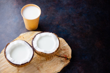 Coconut milk and coconut in paper glass on dark background. Coconut vegan milk non dairy on a wooden stand. Healthy drink concept. Alternative milk. Top view