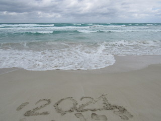 Happy New Year 2021, lettering on the beach with wave and clear blue sea. Numbers 2021 year on the sea shore, message handwritten in the golden sand on beautiful beach background. New Years concept.